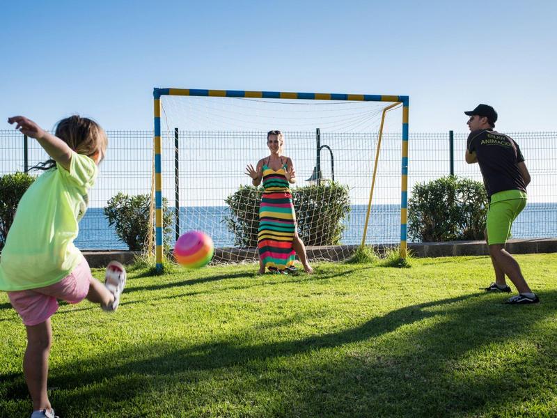 Drei Kinder spielen mit buntem Ball auf grünem Rasen vor blauem Meer und klarem Himmel.