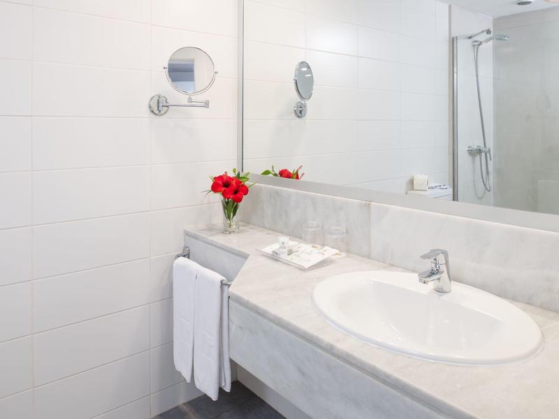 Helles Badezimmer mit großem Spiegel, weißem Waschbecken, Handtüchern und roter Blume in Vase.