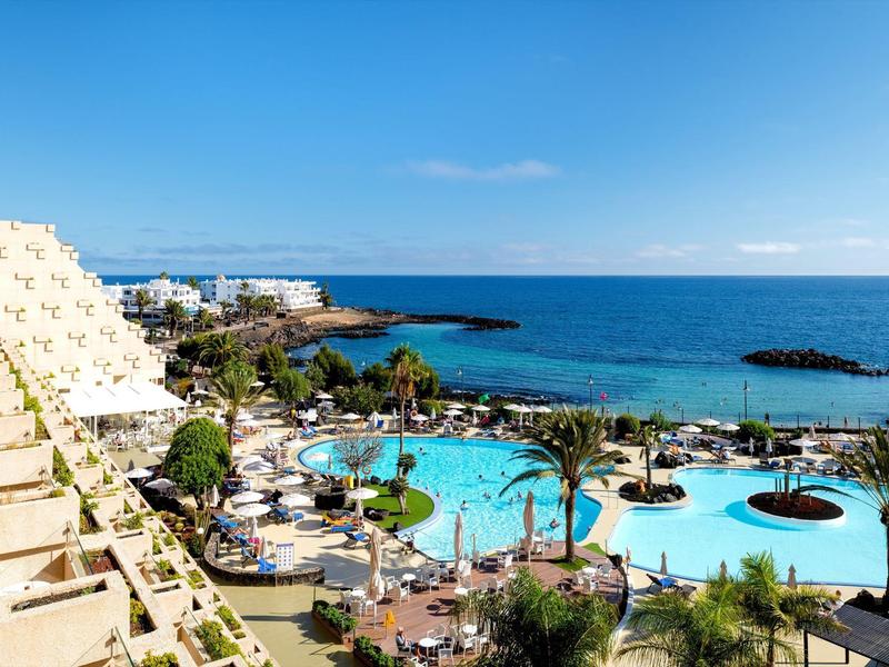 Hotel mit mehreren Pools, Palmen und Meerblick unter klarem blauen Himmel.