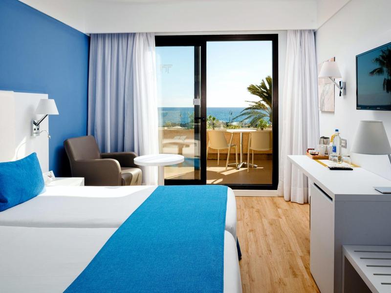 Helles Hotelzimmer mit blau-weißer Einrichtung, Meerblick und Balkon durch große Glasschiebetür.