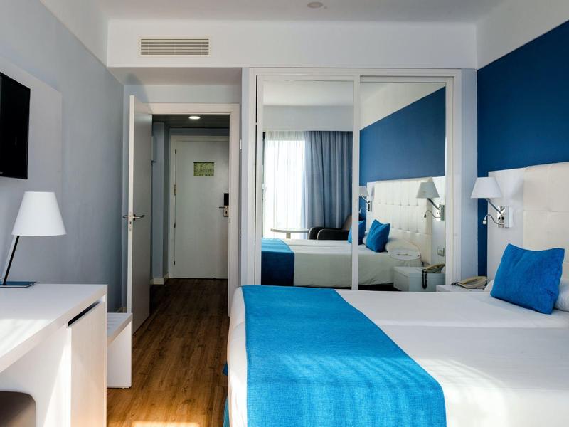 Modernes Hotelzimmer mit Doppelbett, blauen Akzenten und Holzfußboden, seitlich Spiegel und Schreibtisch.