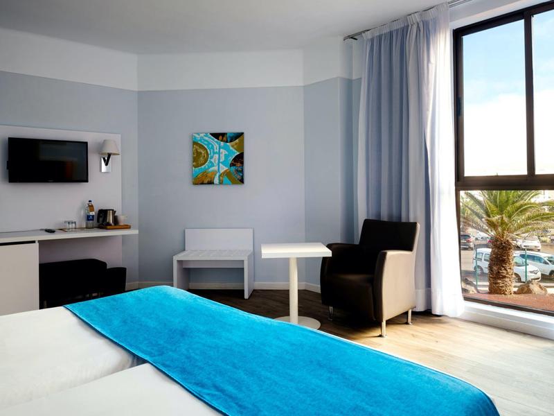 Hell eingerichtetes Hotelzimmer mit Bett, blauem Überwurf, Sesseln, Tisch und großem Fenster.