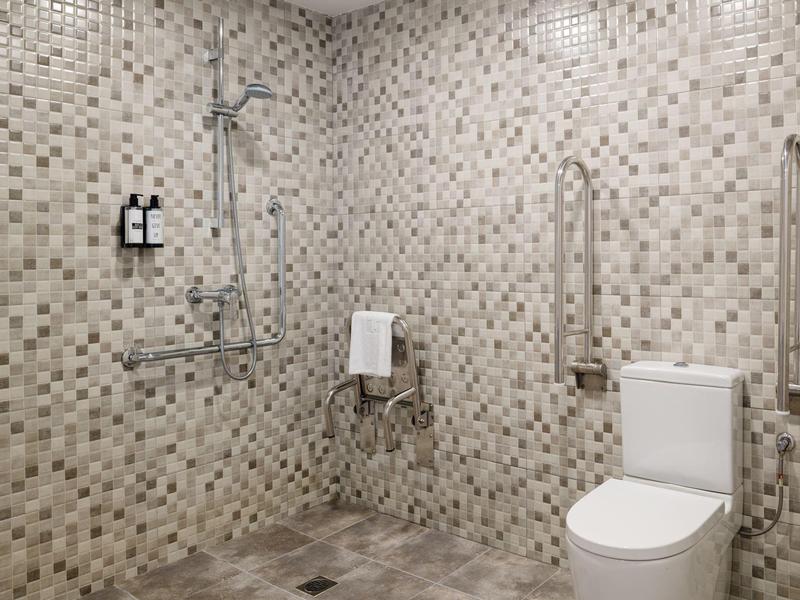 Barrierefreies Badezimmer mit Mosaikfliesen, Dusche mit Haltegriffen und WC mit Stützgriffen.