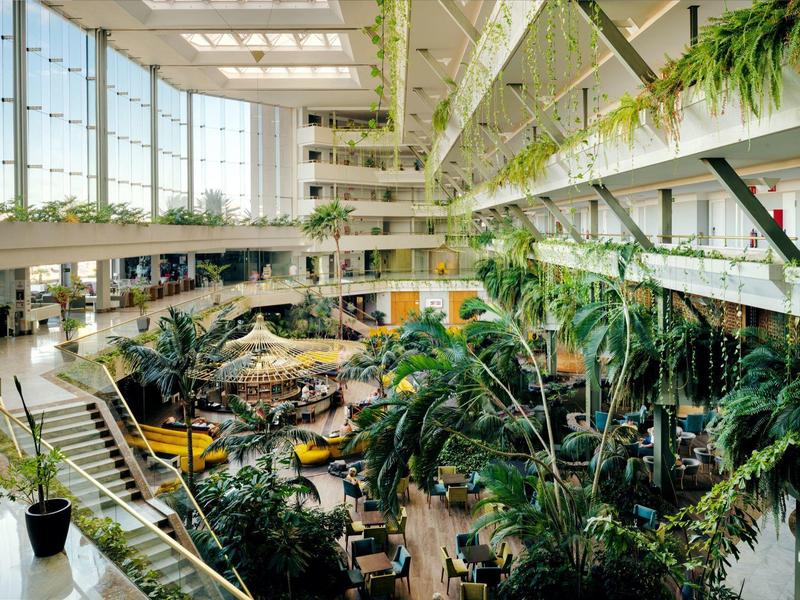 Großes Atrium mit vielen grünen Pflanzen, Glaskuppel und mehrstöckigen Galerien.