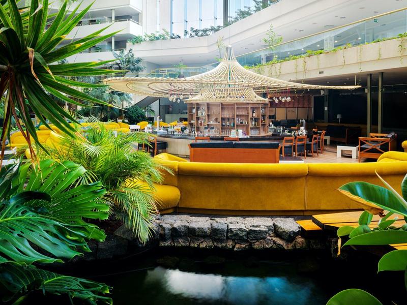 Moderne Bar mit gelben Sitzbänken, grünem Pflanzenarrangement und Wassergraben im Innenbereich.