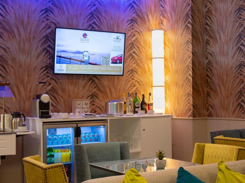 Moderner Barbereich mit TV, Kühlschrank, gelben Sesseln und gestreifter Tapete in warmen Farben.