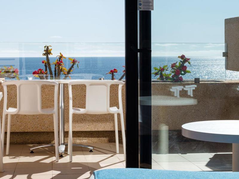 Blick durch Glasschiebetür auf weißen Balkon mit Stühlen und Blumen, im Vordergrund blaue Decke.