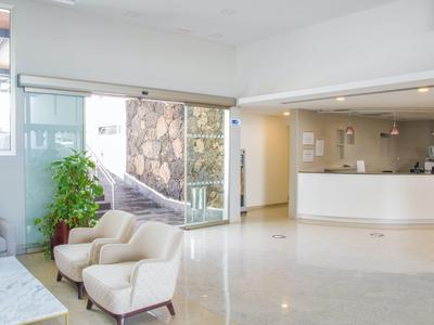 Helle Hotellobby mit weißem Empfangstresen, zwei hellen Sesseln und großem Fenster mit Pflanzen.