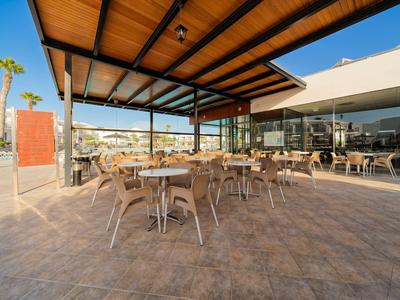Überdachte Terrasse mit Tischen und Stühlen vor einem Hotel bei klarem Himmel.