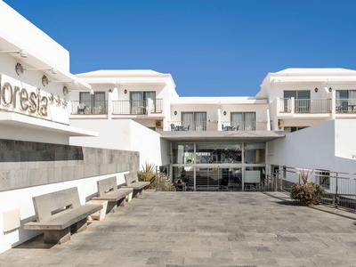 Ingresso principale di un hotel moderno con pareti bianche e panchine in pietra sotto un cielo azzurro.