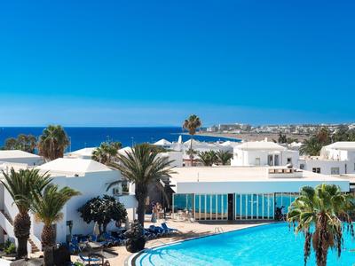 Vista di un hotel bianco con una grande piscina e palme sotto un cielo azzurro.