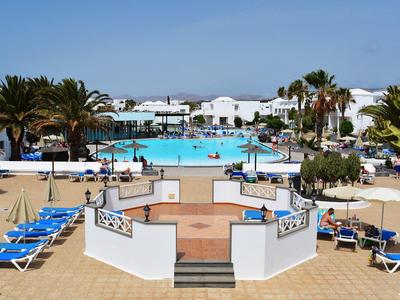 Piscina dell'hotel circondata da palme, lettini blu, ombrelloni e edifici bianchi sotto un cielo azzurro