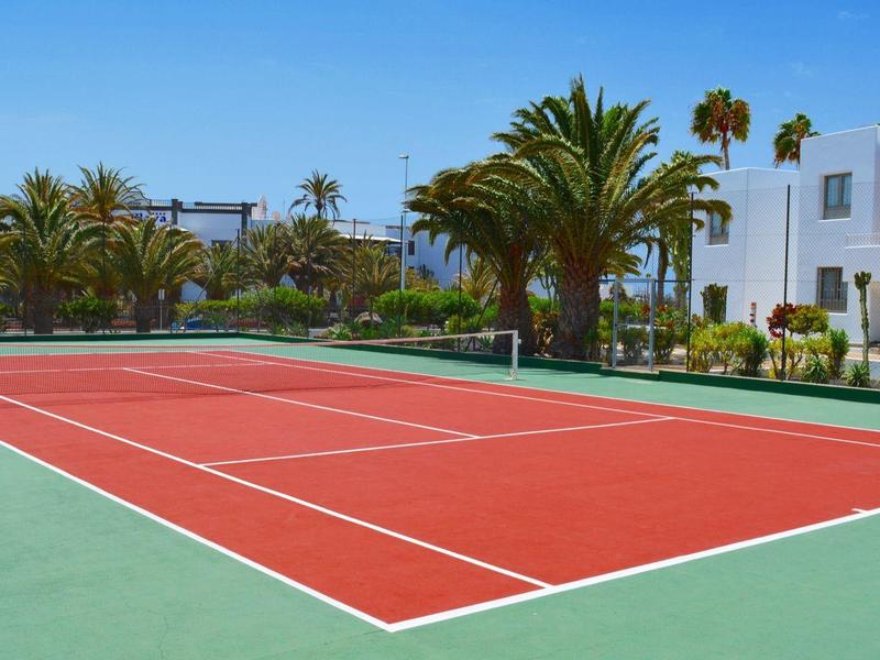 Campo da tennis rosso con palme e edifici bianchi sullo sfondo sotto un cielo azzurro.