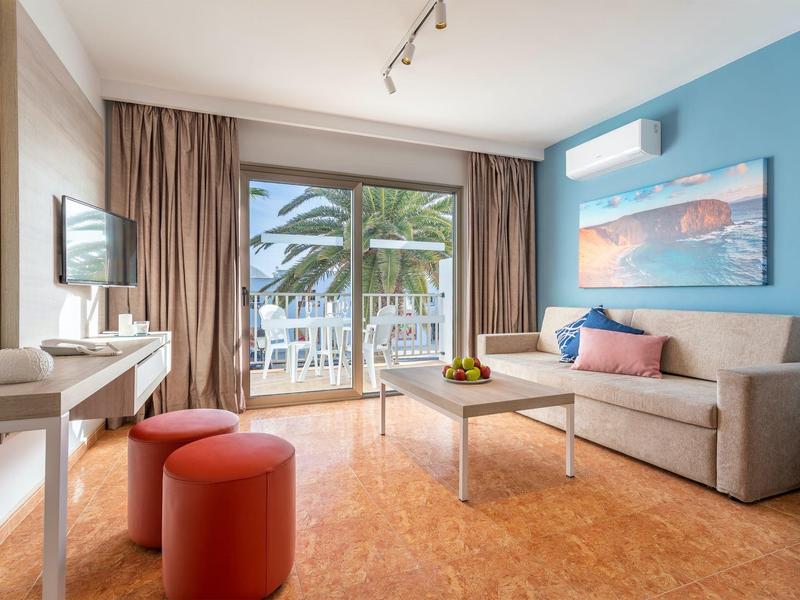 Soggiorno d'hotel moderno con divano grigio, tavolino, pouf rossi e vista su palme.