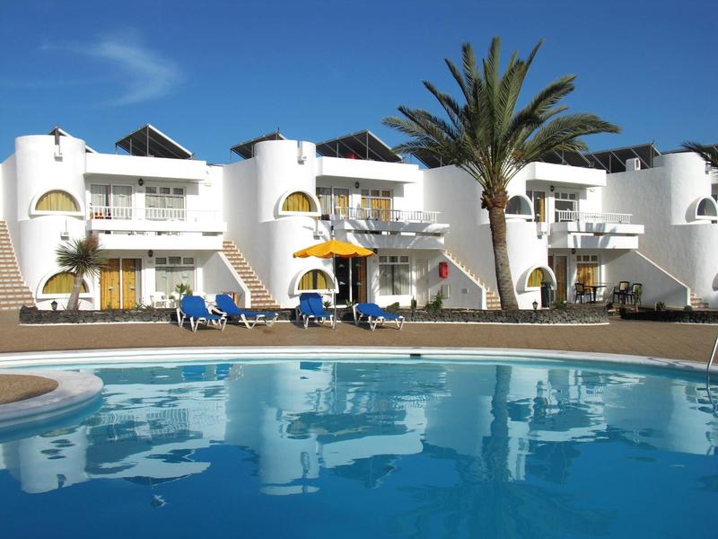 Hotel bianco con piscina, palme, sdraio e ombrelloni gialli sotto un cielo azzurro.