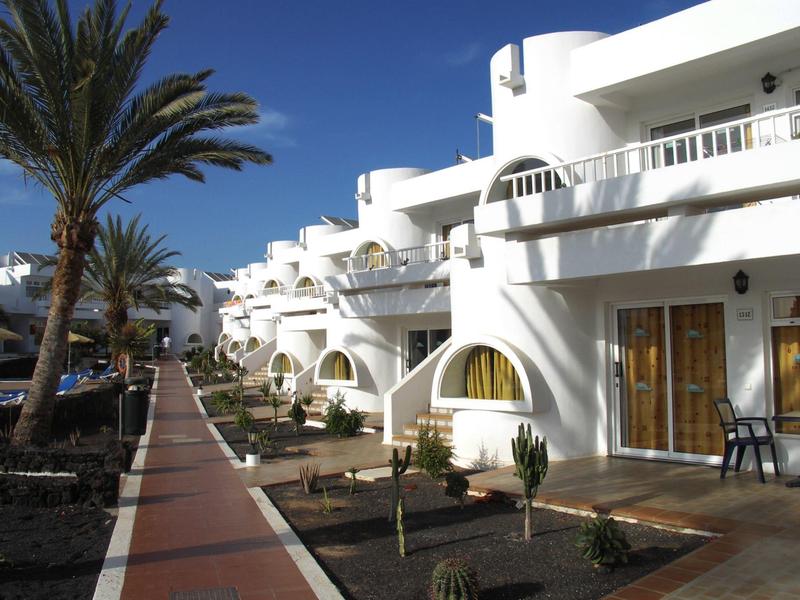 Hotel bianco con balconi e palme lungo una passerella lastricata.