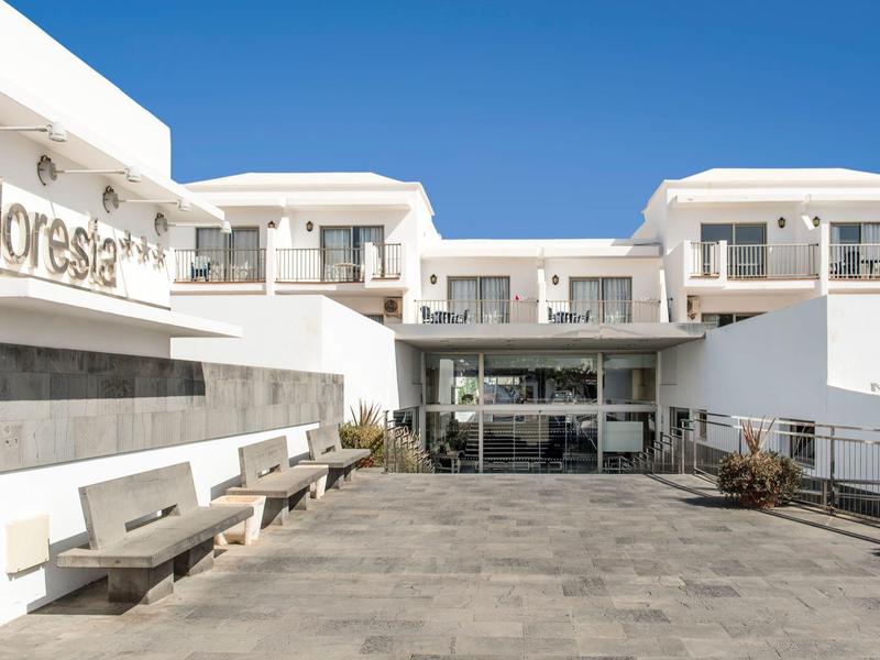 Ingresso principale di un hotel moderno con pareti bianche e panchine in pietra sotto un cielo azzurro.