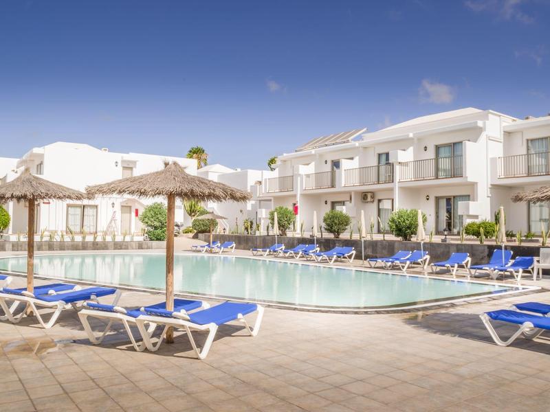 Piscina all'aperto con lettini e ombrelloni, hotel moderni sullo sfondo sotto cielo azzurro.
