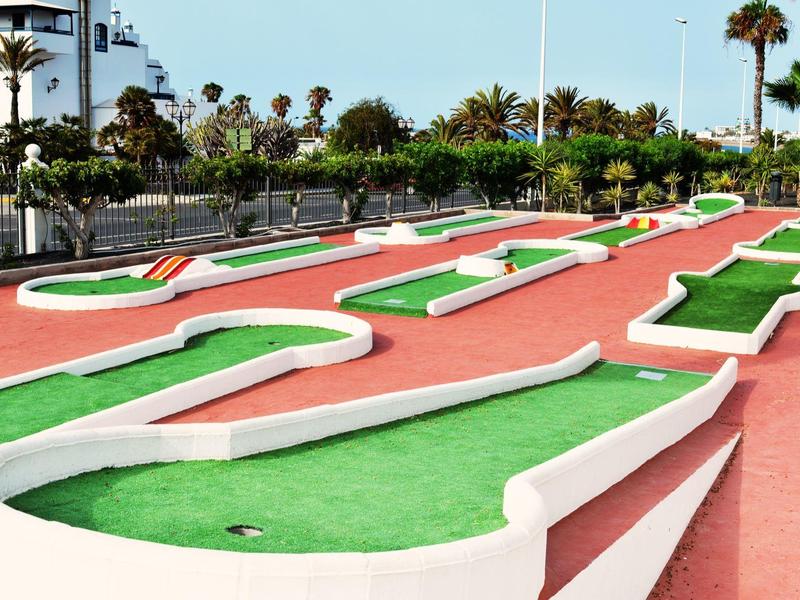 Percorso di mini golf con ostacoli bianchi e superficie verde, circondato da palme.