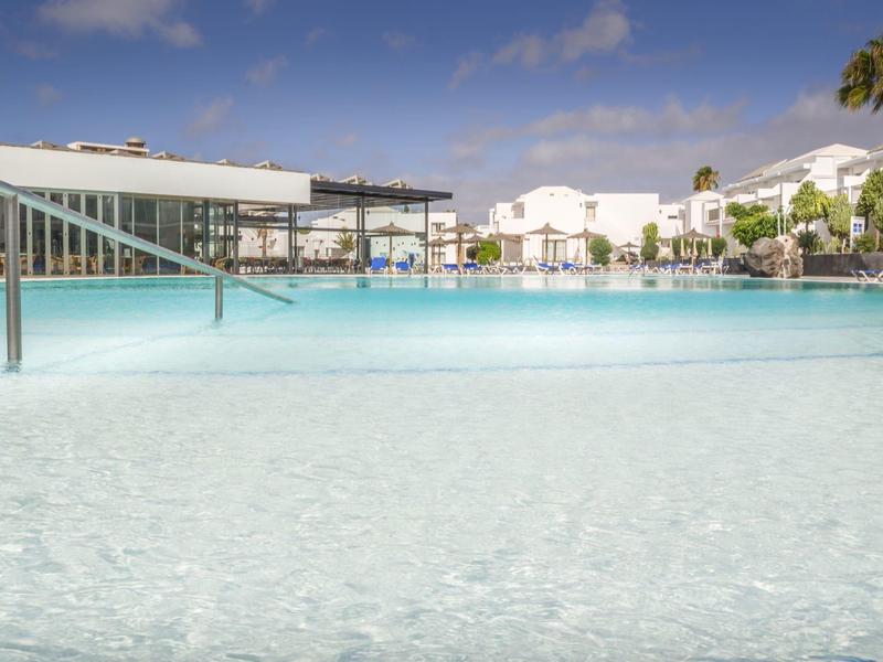 Grande piscina limpida con edifici bianchi e palme sotto un cielo azzurro.