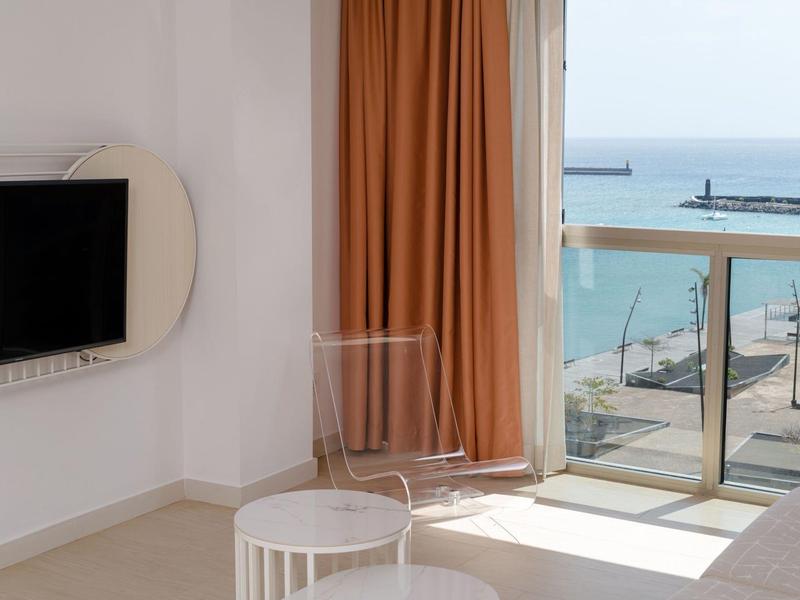 Helles Hotelzimmer mit Fernseher, orangenen Vorhängen und Meerblick durch die Balkontür.