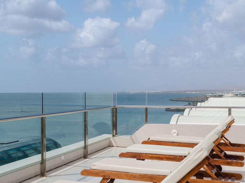 Mehrere Liegestühle auf Balkon mit Glasgeländer und Meerblick bei bewölktem Himmel.