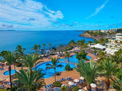 Duży basen hotelowy z palmami i widokiem na morze w słoneczny dzień.