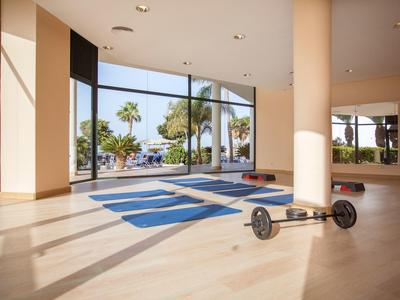 Jasna siłownia z niebieskimi matami do jogi i sztangą przed dużymi oknami z widokiem na palmy.