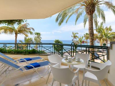 Balkon s květináči, posezením a dvěma lehátky s výhledem na tropické palmy a moře.