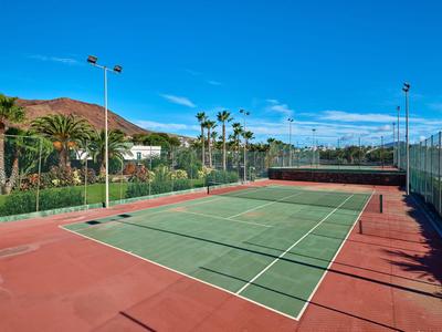 Venkovní tenisový kurt s červenými a zelenými plochami, obklopený palmami a modrou oblohou.