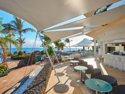 Otwarta przestrzeń restauracyjna z stołami, żaglami przeciwsłonecznymi i widokiem na morze i palmy.