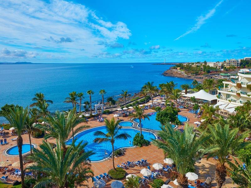 Duży basen hotelowy z palmami i widokiem na morze w słoneczny dzień.