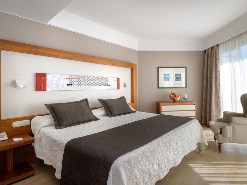 Nowoczesny pokój hotelowy z łóżkiem podwójnym, szarymi poduszkami i fotelem obok okna z zasłonami.