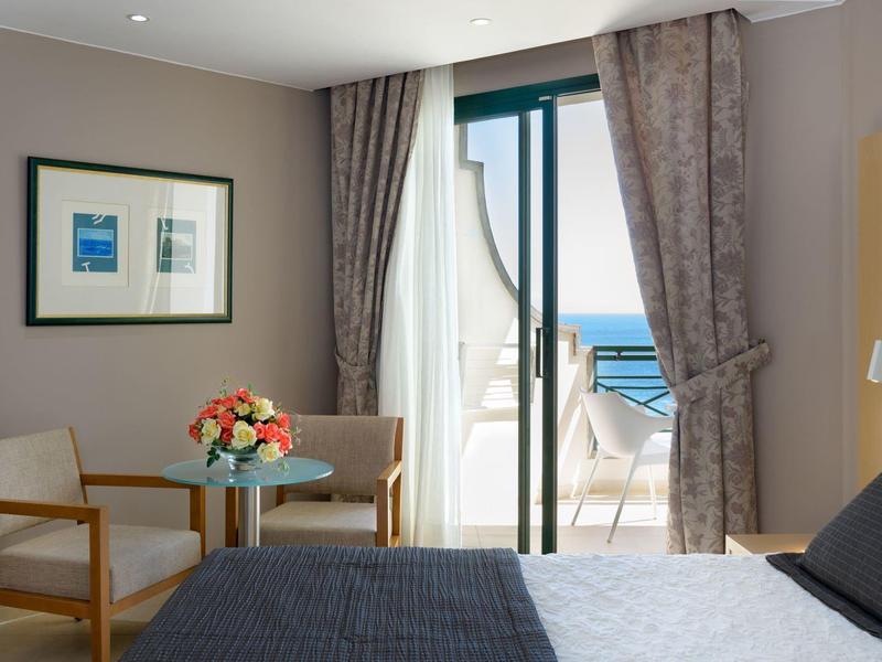 Jasny pokój hotelowy z łóżkiem, stołem, krzesłami i balkonem z widokiem na morze.