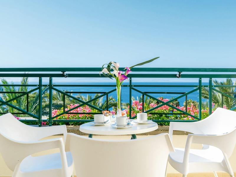 Dwa białe krzesła i stół z kwiatami z widokiem na morze na tarasie hotelowym.