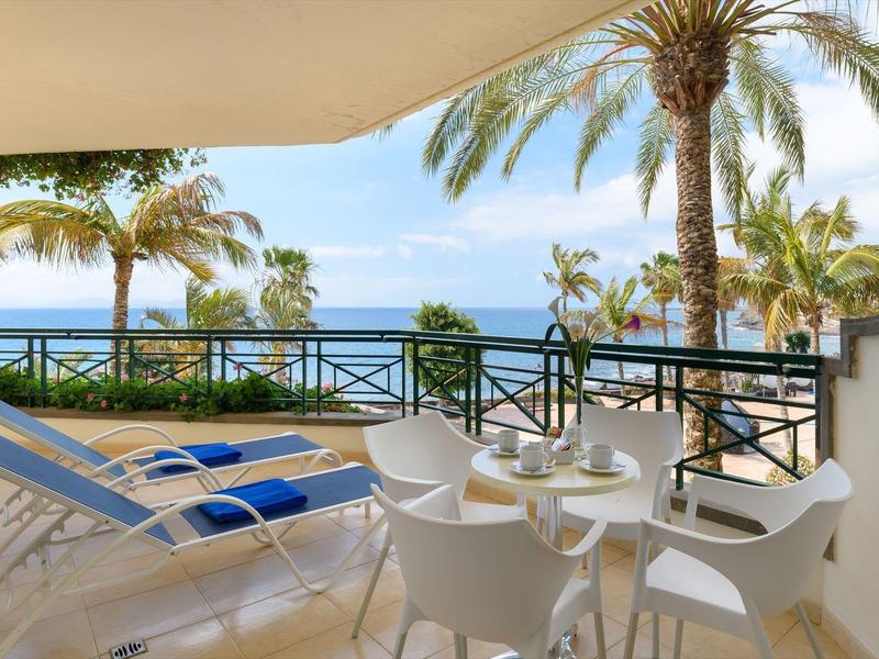 Balkon z białymi krzesłami i niebieskimi leżakami z widokiem na morze i palmy.