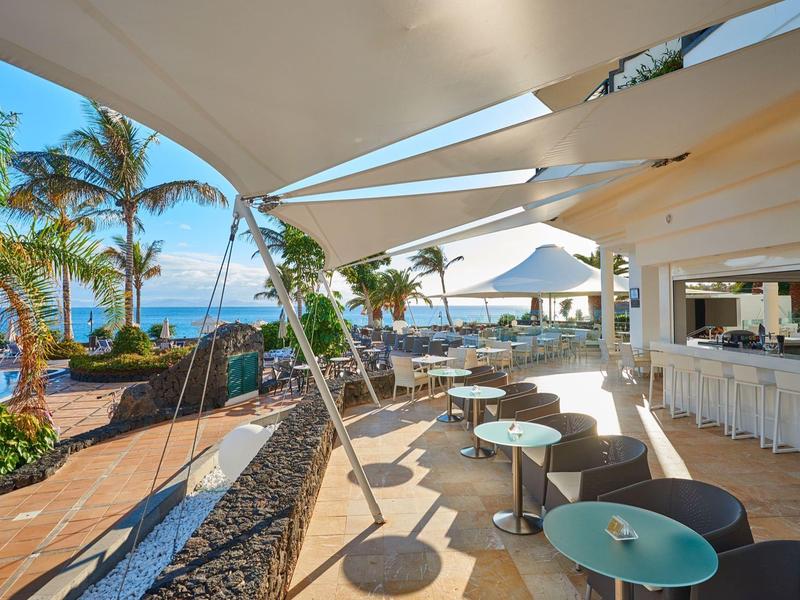 Otwarta przestrzeń restauracyjna z stołami, żaglami przeciwsłonecznymi i widokiem na morze i palmy.