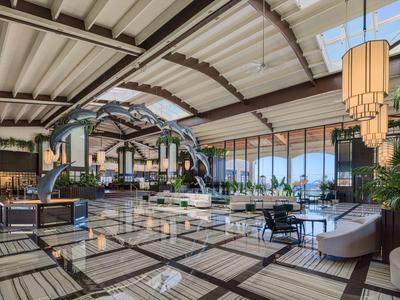 Spacious hotel lobby with modern furniture, large windows, and an airy, geometric ceiling design.