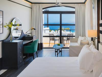 Bright hotel room with bed, desk, sofa, and balcony overlooking ocean and beach.