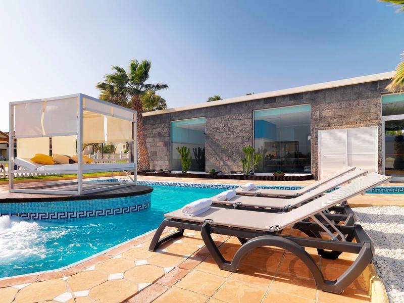 Modern hotel pool area with loungers, cabana, and a sleek building under a clear sky.