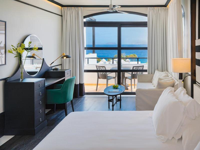 Bright hotel room with bed, desk, sofa, and balcony overlooking ocean and beach.