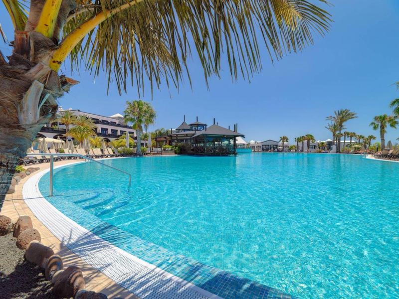 Resort pool with clear blue water, palm trees, and a curved edge under a sunny sky.