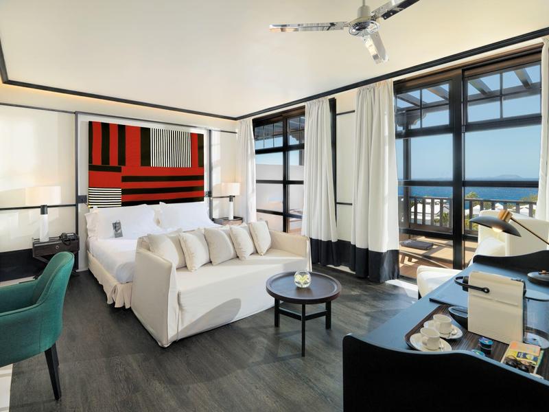 Luxurious hotel room with white bed, modern decor, balcony overlooking sea, and elegant seating area.