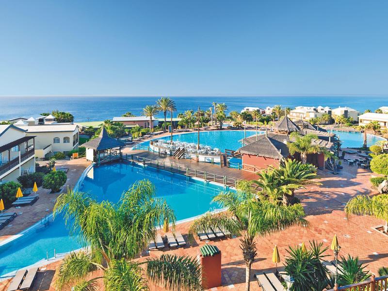 Resort pool area with lounge chairs, palm trees, and ocean view under clear blue sky.