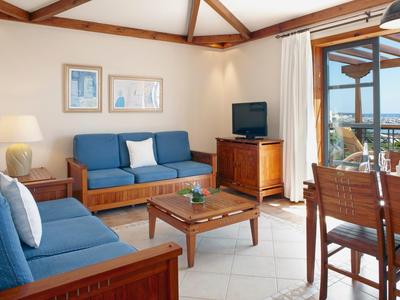 Helles Wohnzimmer mit Holzmöbeln, blauen Sofas, Fernseher und Terrassentür mit Meerblick.