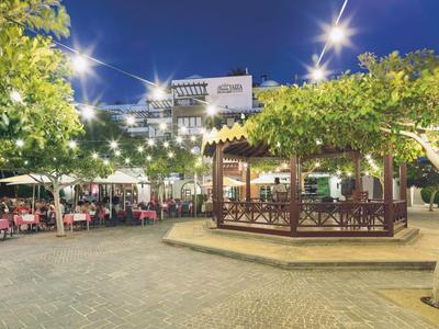 Abendliche Straßenszene mit beleuchtetem Pavillon, Tischen, Stühlen und Bäumen unter blauem Himmel.