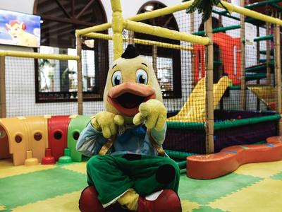 Ein Enten-Maskottchen sitzt vor einem bunten Indoor-Spielplatz mit Klettergerüsten.