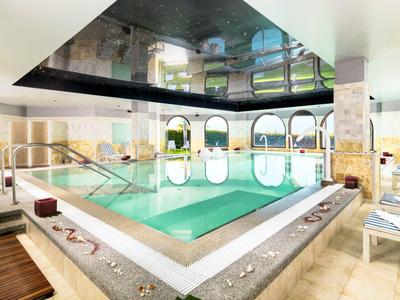 Moderner Innenpool mit heller Marmorumgebung, Holzliege und Spiegeldecke, große Fenster lassen Licht herein.