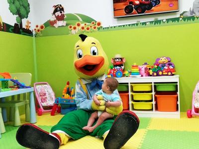 Bunter Spielraum mit grünem Boden und Wänden, großem Entenmaskottchen und Kleinkind.
