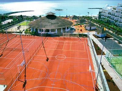 Mehrere rote Tennisplätze mit Zäunen, daneben weiße Gebäude und Meer im Hintergrund.